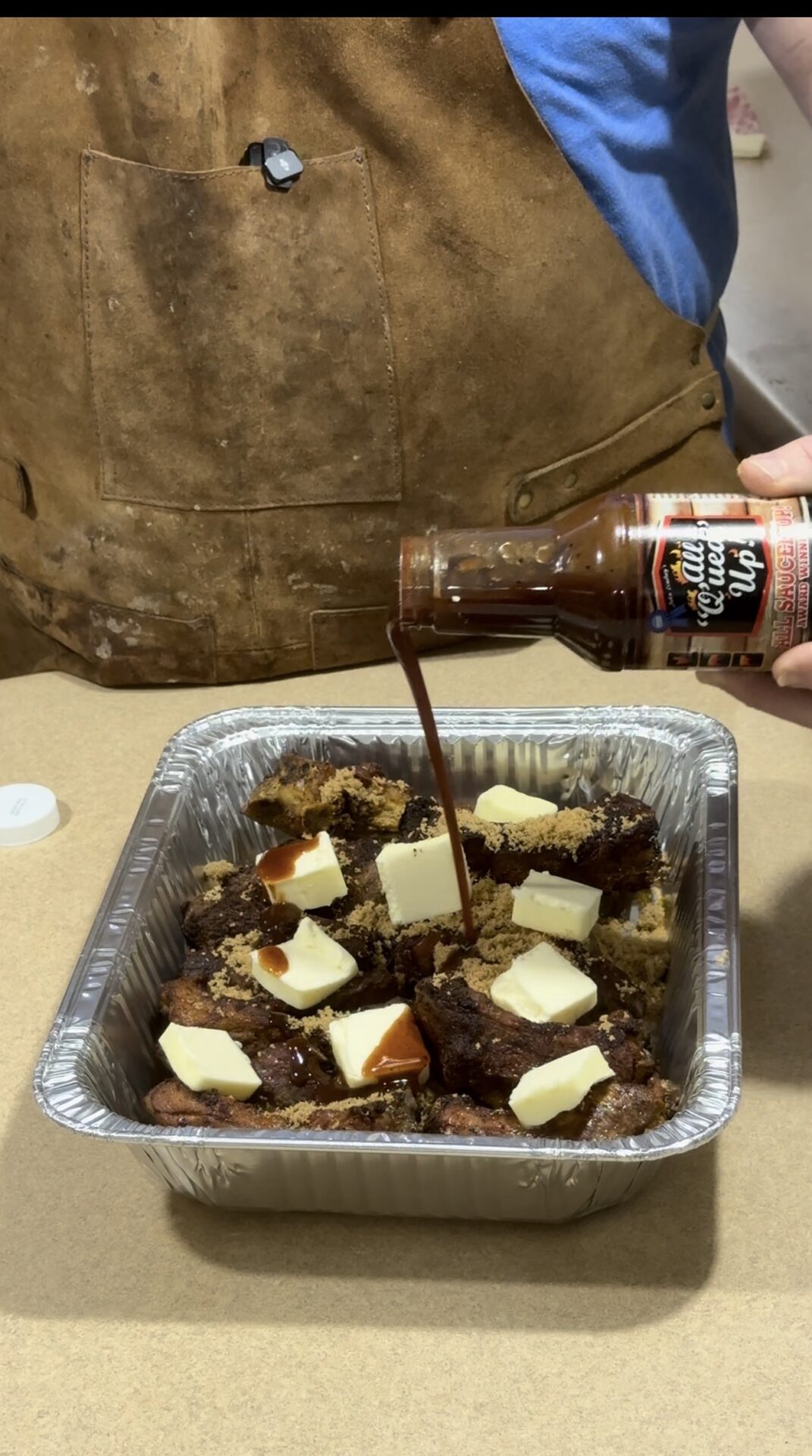 A person pours barbecue sauce from a bottle over beef short ribs in a foil tray. The ribs are topped with cubes of butter and brown sugar. The person wears a brown apron and a blue shirt.