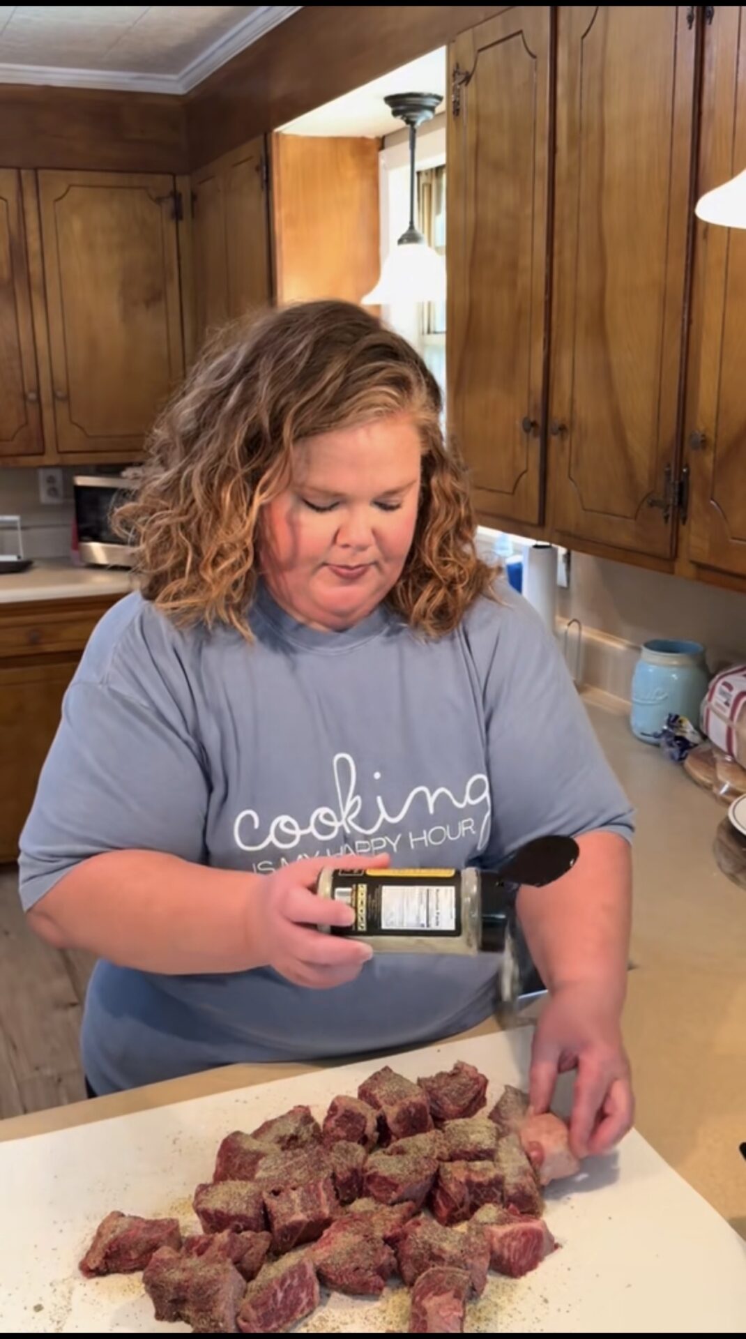 A woman with shoulder-length curly hair seasons chunks of raw meat on a kitchen counter. She is wearing a light blue shirt that says cooking is my happy hour. Wooden cabinets and kitchen appliances are visible in the background.