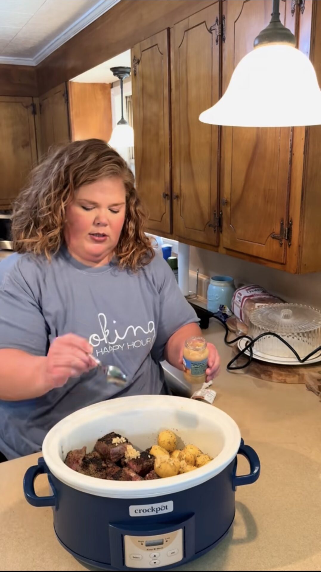 A woman stands in a kitchen adding seasoning from a jar into a Crockpot filled with meat and potatoes on the counter. Wooden cabinets and kitchen appliances are visible in the background.