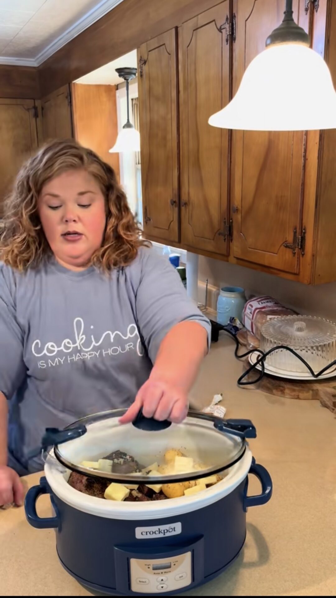 A woman in a light blue Cooking is my happy hour shirt is using a blue Crockpot slow cooker in a kitchen with wooden cabinets and tan countertops under warm hanging lights.