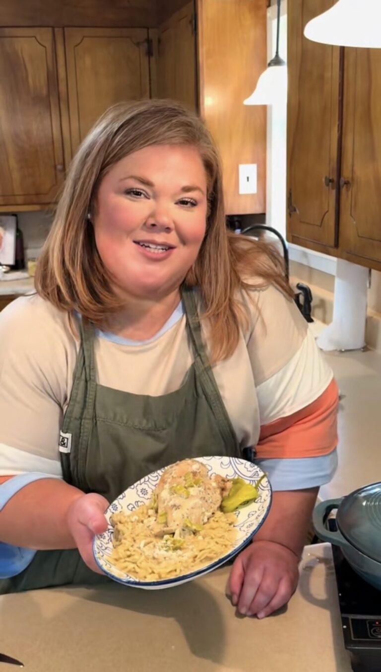 A woman in an apron stands in a kitchen, smiling and holding a plate of rice and chicken with vegetables toward the camera. Wooden cabinets and countertops are visible in the background.
