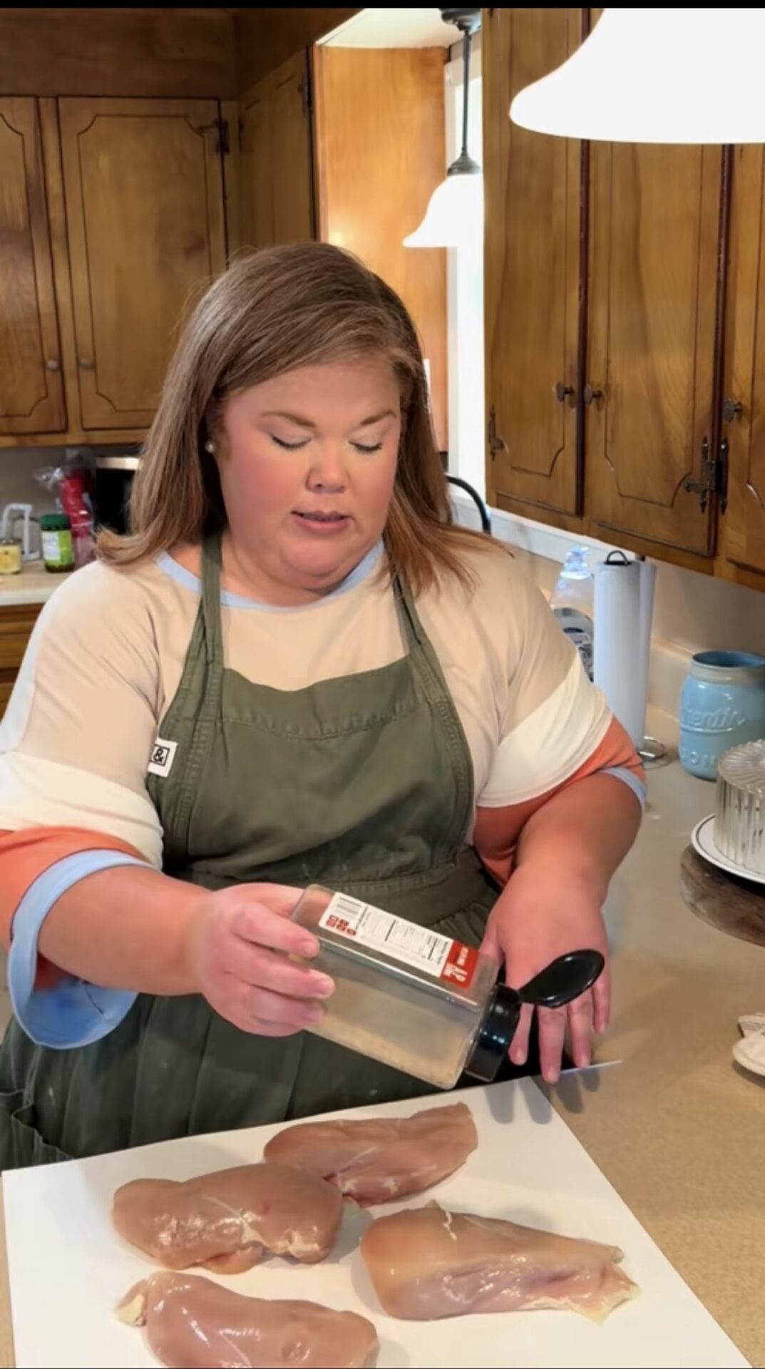 A woman in a green apron seasons raw chicken breasts on a kitchen counter with a shaker of spices. Wooden cabinets and kitchen items are visible in the background.