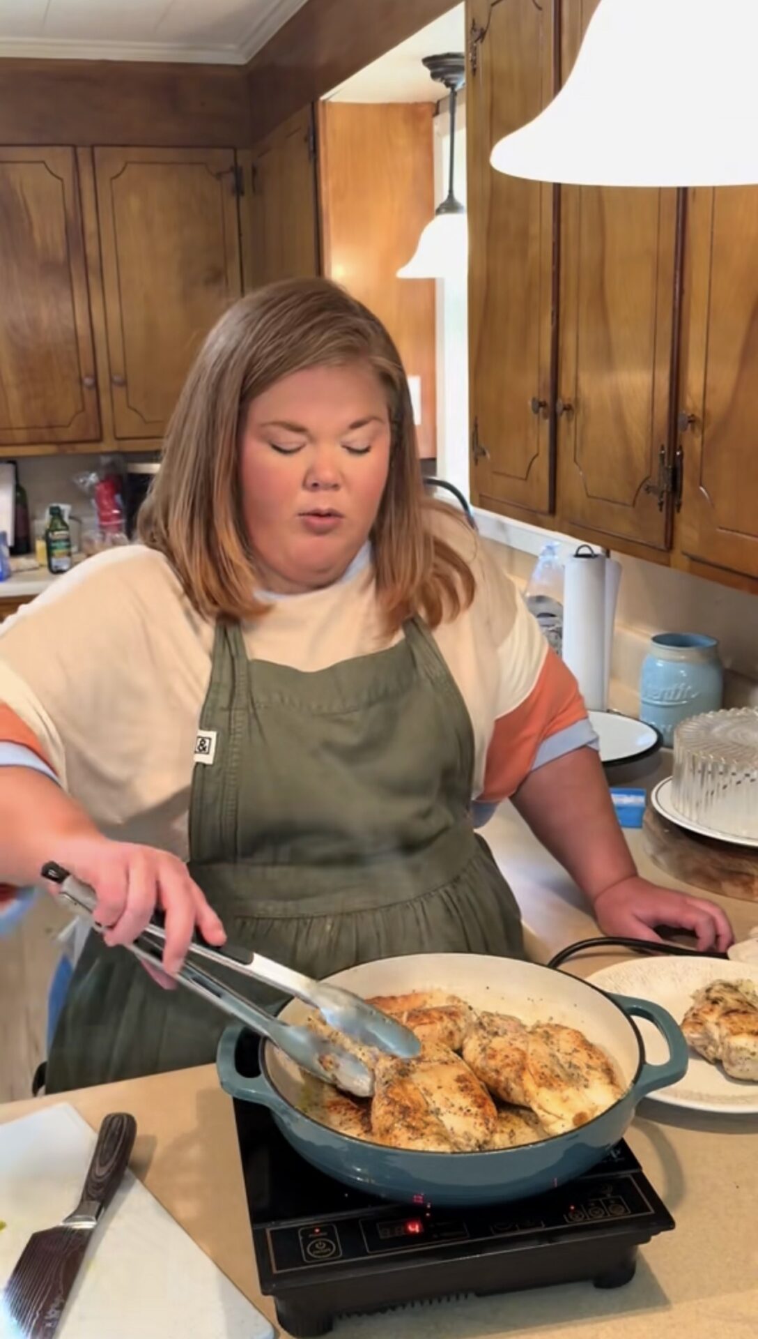 A woman wearing a green apron cooks seasoned chicken in a pan on a stovetop in a kitchen, using tongs to turn the pieces. Wooden cabinets and kitchen items are visible in the background.