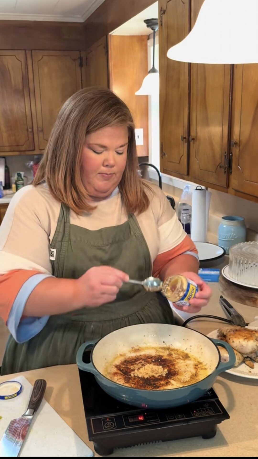 A woman wearing an apron stands in a kitchen, holding a spoonful of minced garlic over a pan on a stovetop. Food and cooking utensils are on the counter, and wooden cabinets are visible in the background.