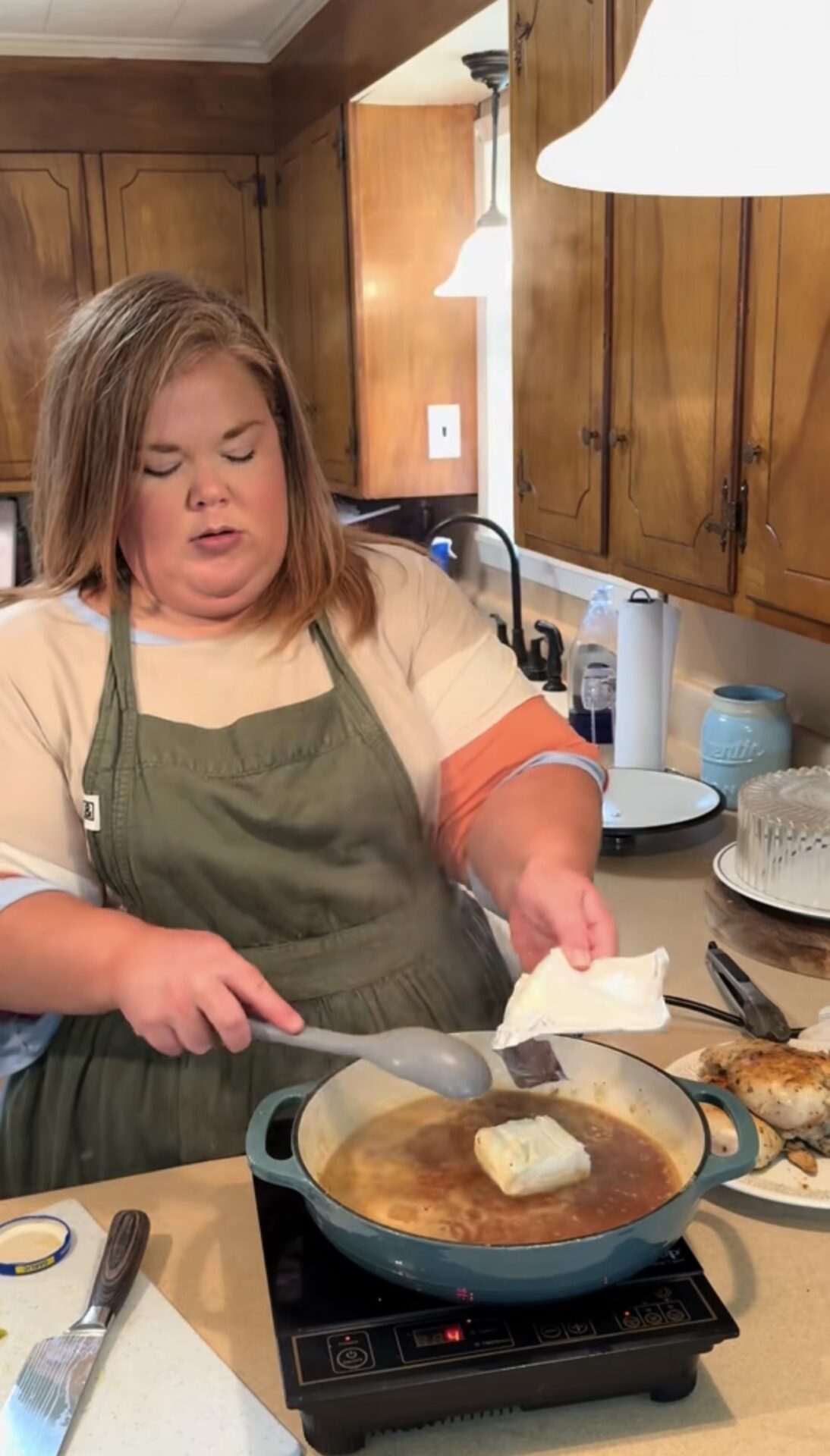 A woman wearing a green apron stirs a brown sauce in a pan on an electric stove, preparing to add a block of cream cheese. She is in a kitchen with wooden cabinets and various utensils on the counter.