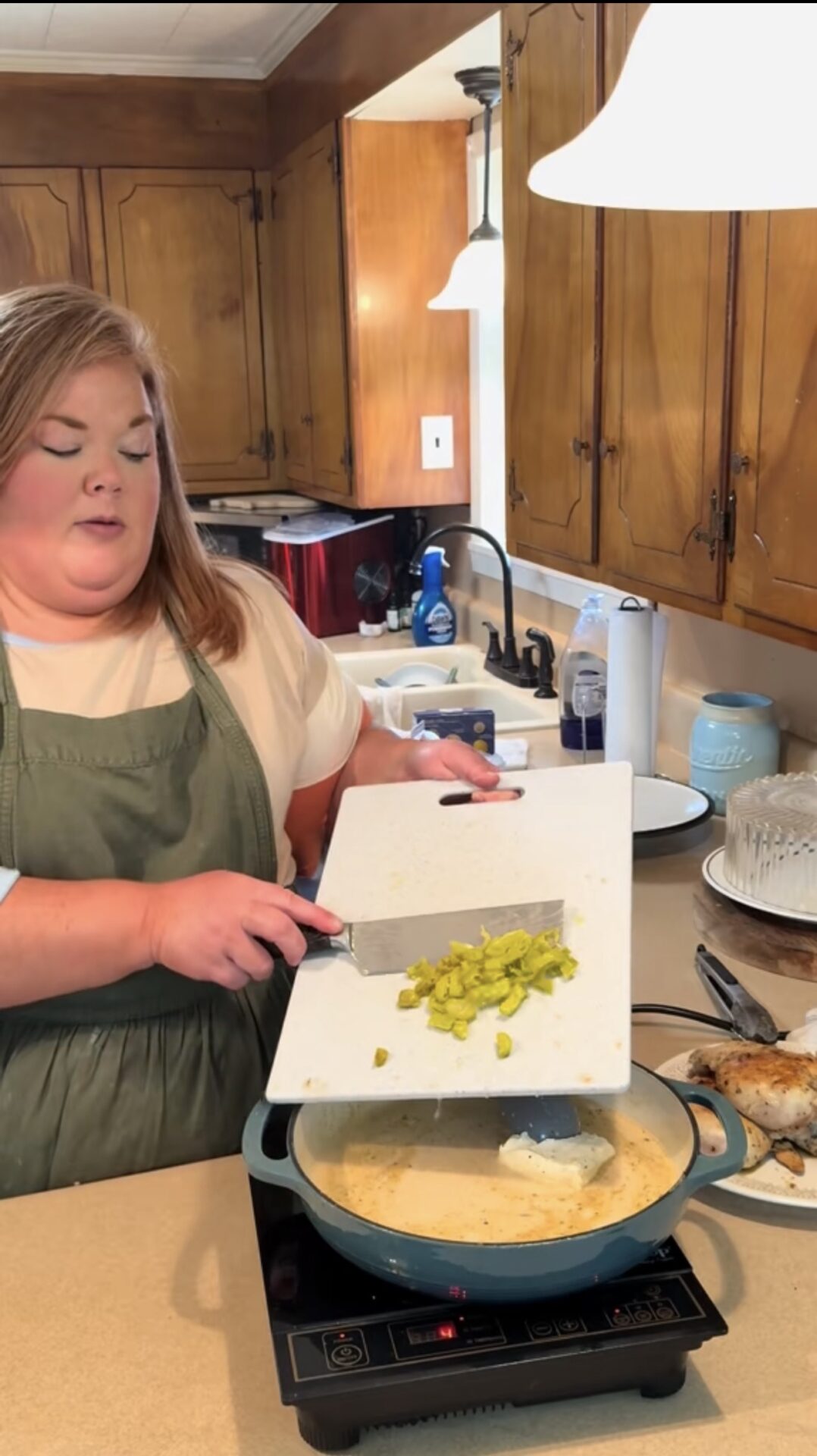 A woman in a green apron stands in a kitchen, adding chopped green vegetables from a cutting board into a pan of creamy sauce on an induction cooktop. Cooked chicken sits on a plate beside her.