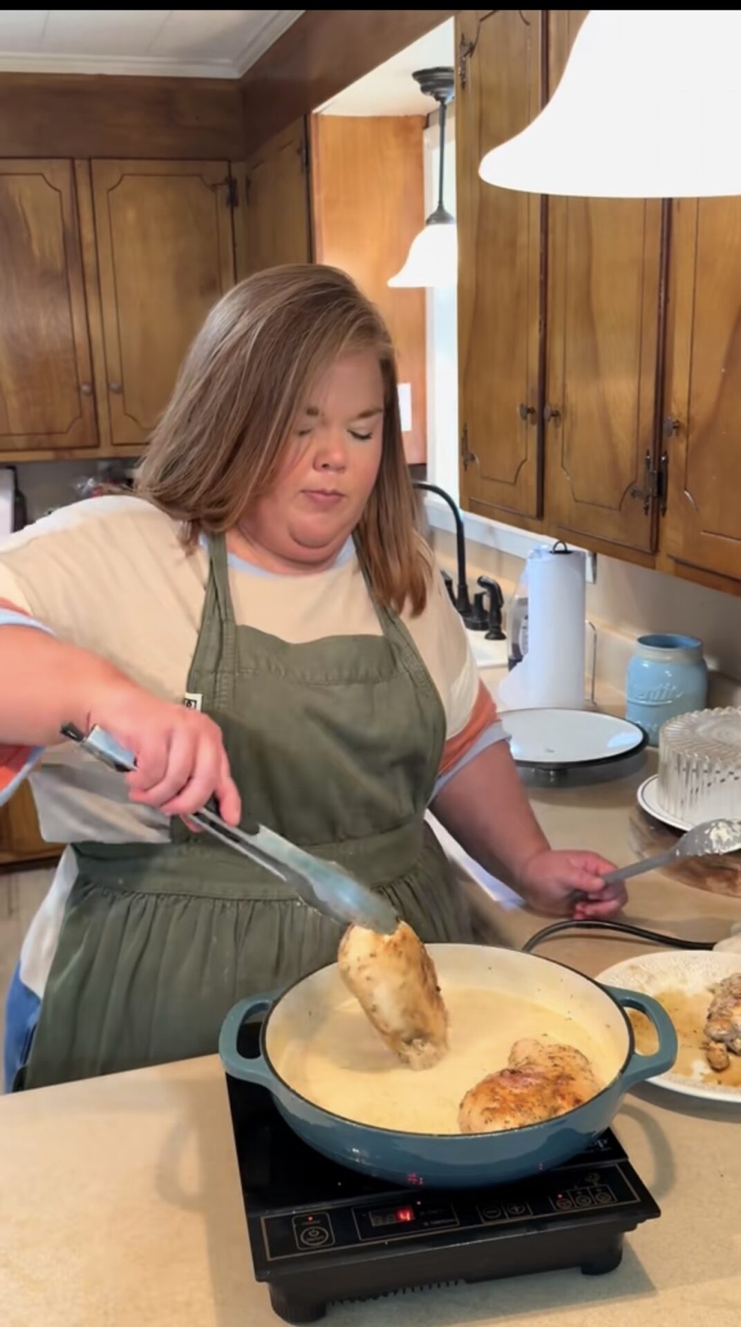 A woman in a green apron cooks chicken in a pan on a stovetop in a kitchen with wooden cabinets. She uses tongs to lift a piece of chicken, and there are plates and kitchen items on the counter.