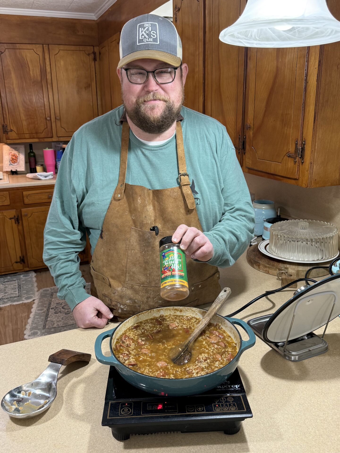 A man in glasses and an apron stands in a kitchen, holding a spice jar above a pot of stew cooking on an electric stove. The kitchen has wooden cabinets and a cake dish on the counter.