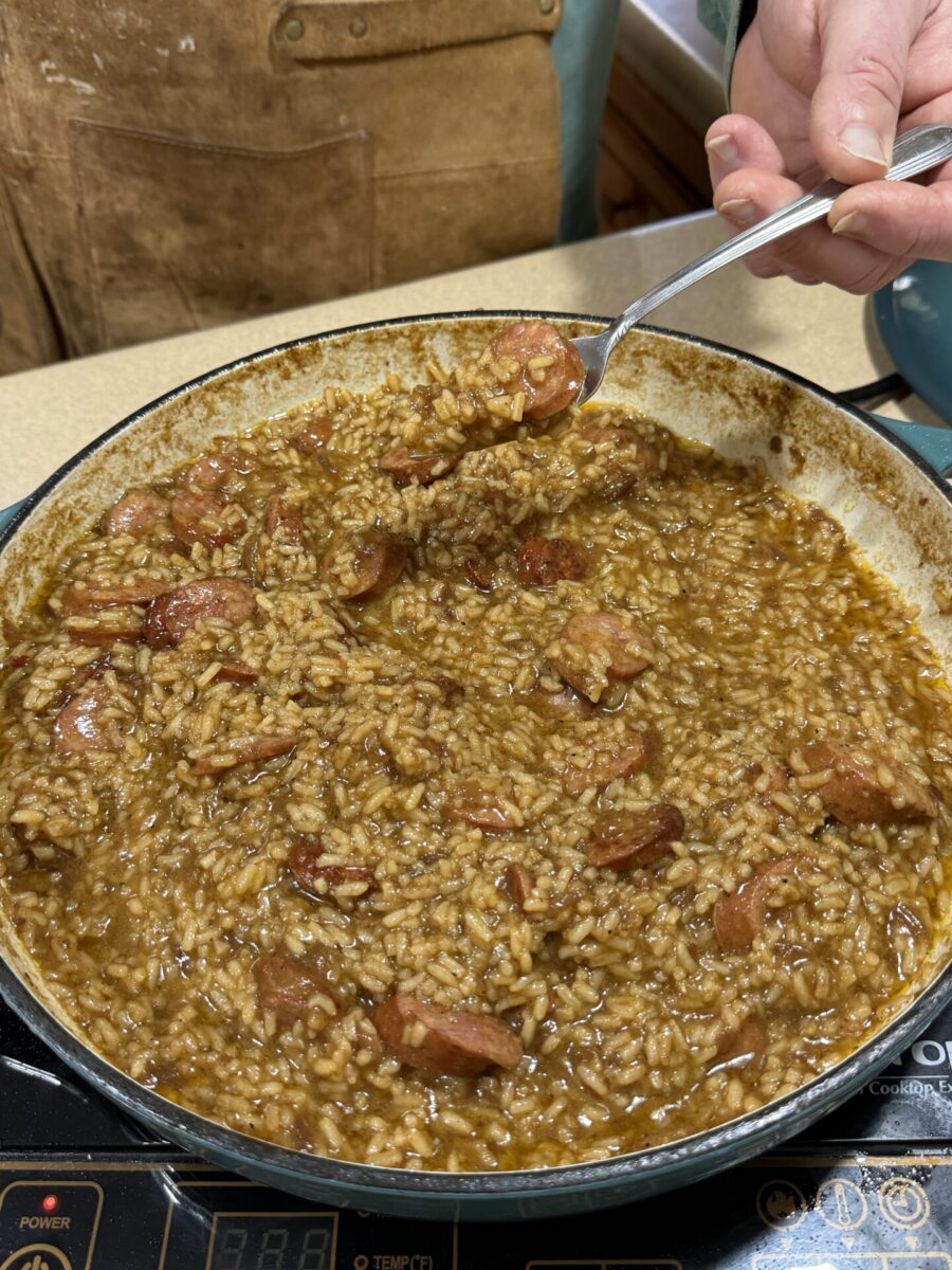 A hand holds a spoonful of sausage and rice from a large skillet filled with a thick, brown, savory mixture, likely a homemade jambalaya or similar dish, on a stovetop.