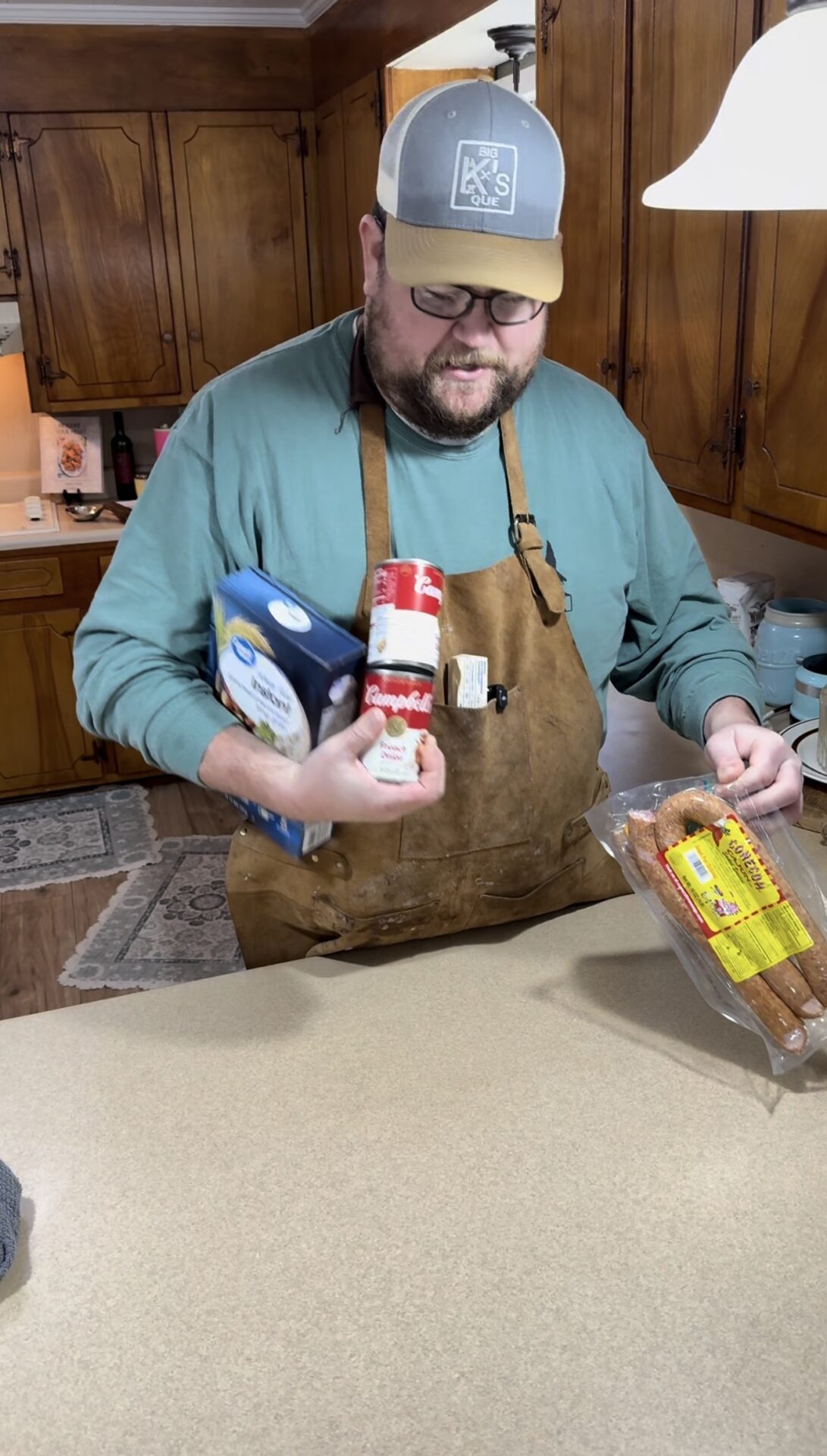 A man wearing a cap, glasses, and an apron stands in a kitchen holding a container of Morton salt, a can of Campbell’s soup, and a package of Hillshire Farm sausage on a counter.