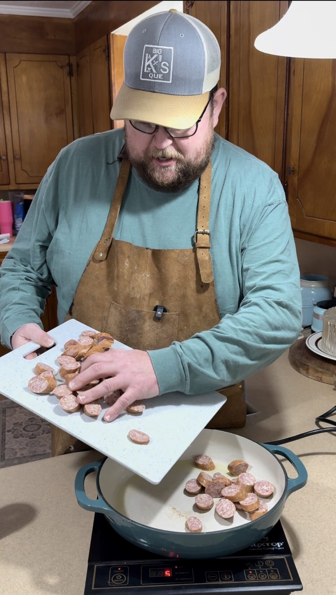 A man wearing a cap, glasses, apron, and long-sleeved shirt adds sliced sausage from a cutting board into a large pan on an electric stove in a kitchen with wooden cabinets.