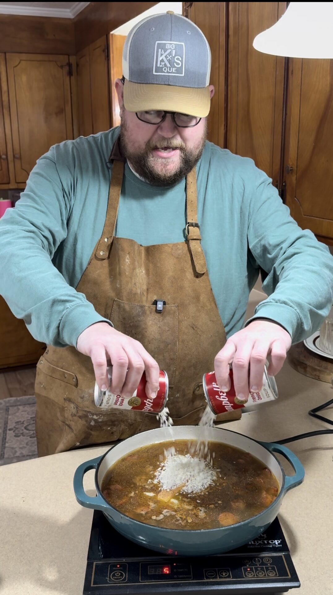 A man in a cap, glasses, and apron stands in a kitchen, pouring the contents of two cans into a pot of soup on an induction cooktop. The kitchen has wooden cabinets and plates are stacked nearby.