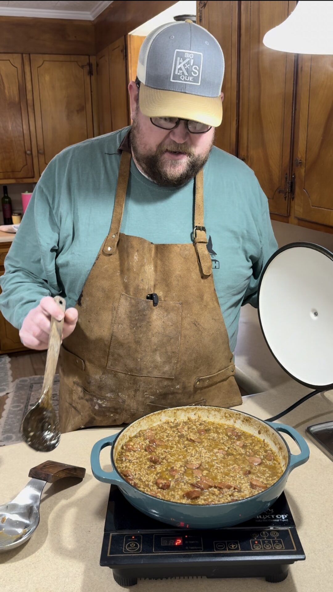 A bearded man in a hat, glasses, and a brown apron stands in a kitchen ladling a hearty stew or gumbo from a large pot on an induction cooktop, holding the lid in his other hand.