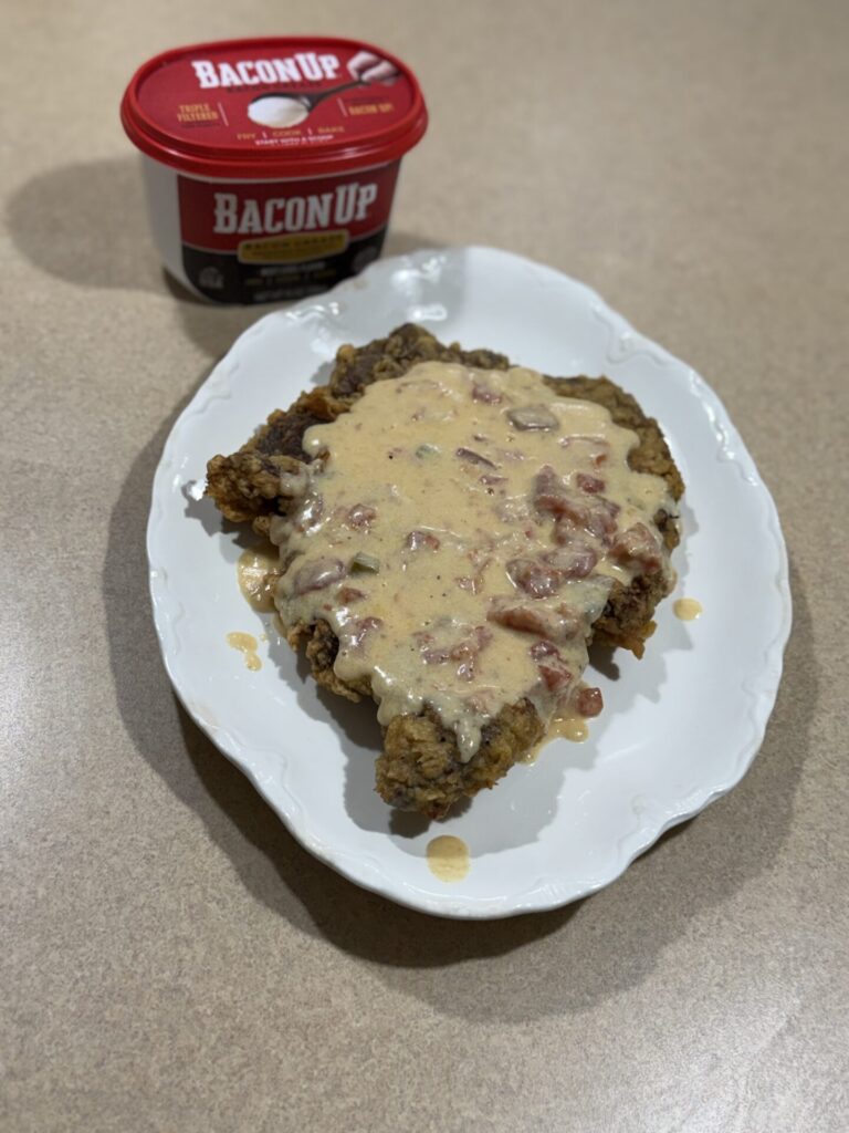 A piece of fried steak is topped with creamy gravy on a white oval plate. Behind the plate is a red tub labeled Bacon Up on a beige countertop.