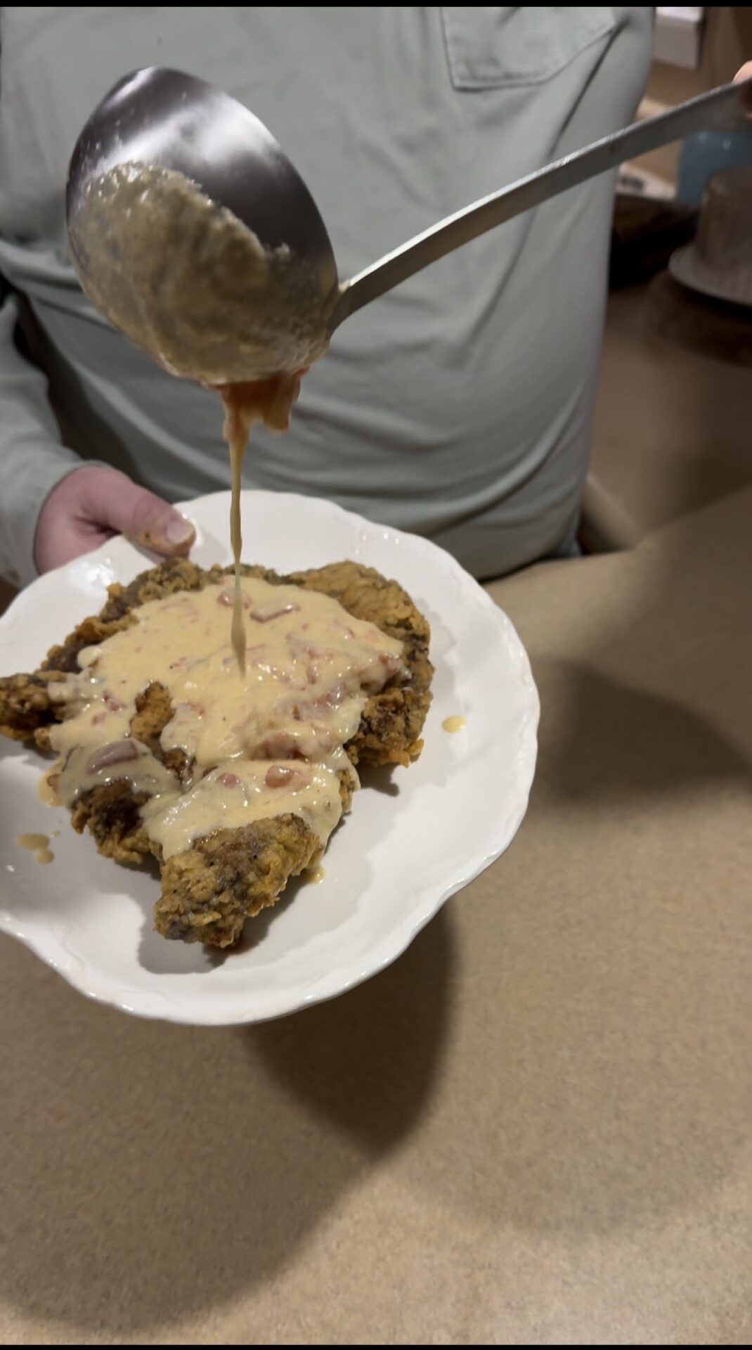 A person pours creamy gravy with bits of tomato over a piece of fried chicken steak on a white plate, standing at a kitchen counter.