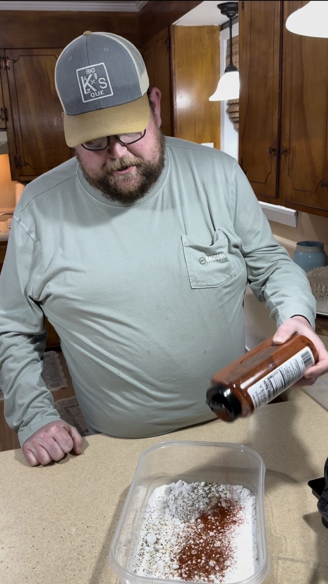 A man in a light green long-sleeve shirt and cap is pouring seasoning from a bottle onto flour in a container on a kitchen counter, with wooden cabinets and warm lighting in the background.