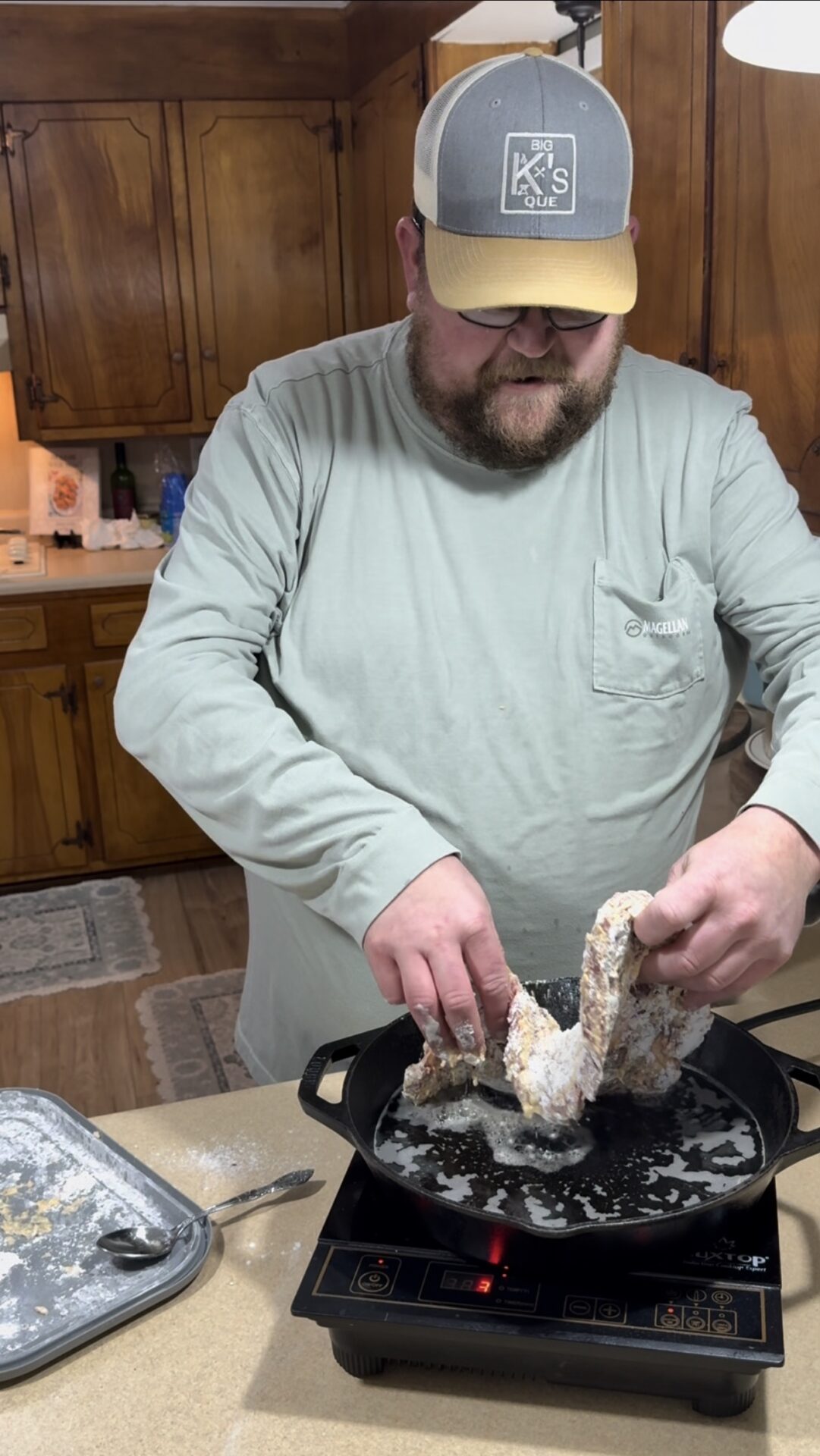 A man in a light green shirt and cap is placing breaded pieces of chicken into a cast iron skillet with oil on a stovetop in a wood-paneled kitchen.