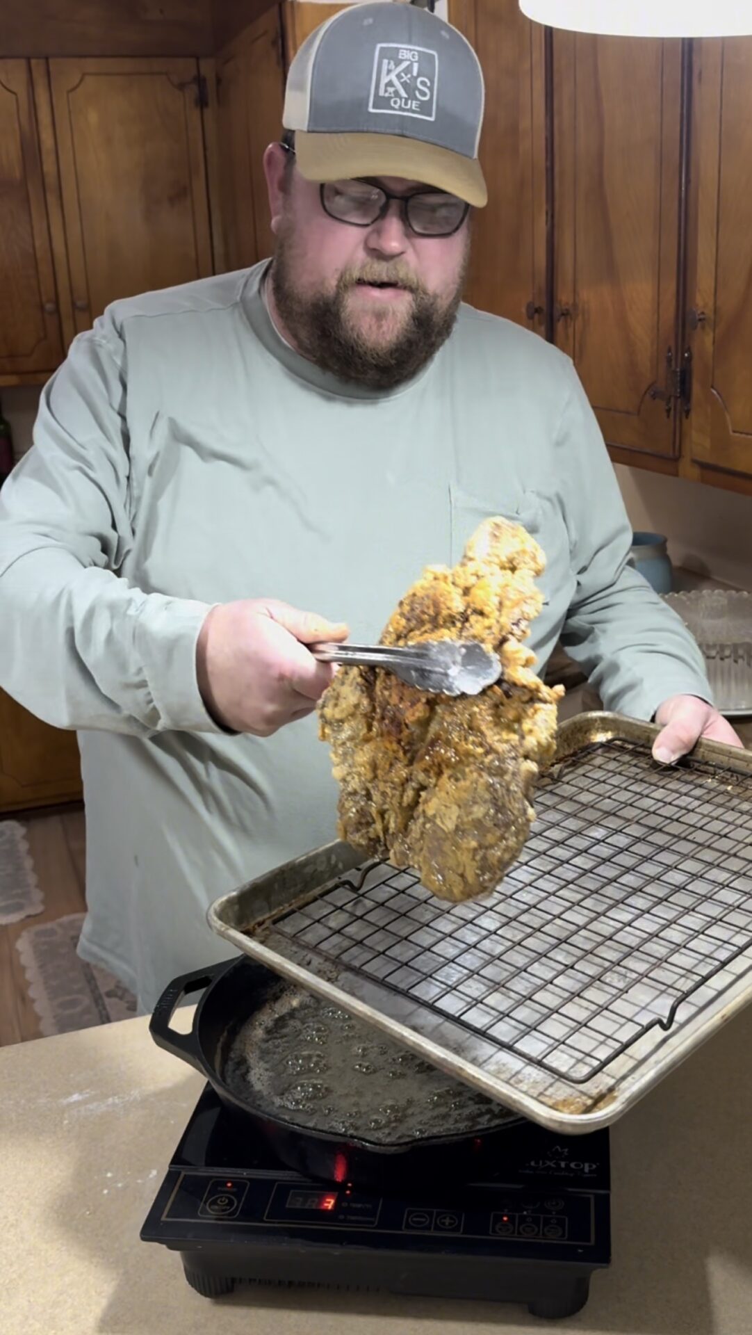 A man in glasses and a hat holds a pair of tongs with a large piece of fried steak over a tray with a wire rack, standing in a kitchen with wooden cabinets.