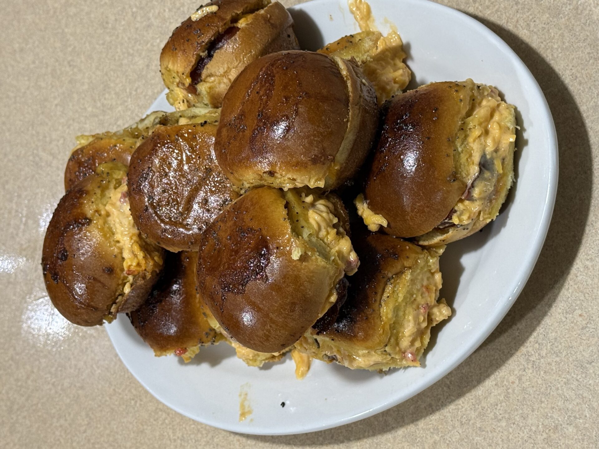 A white plate piled with several small, shiny brown slider buns filled with a creamy, orange-colored mixture, sitting on a beige countertop.