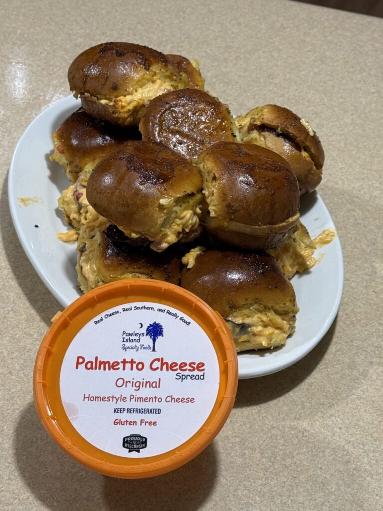 A plate of slider sandwiches filled with pimento cheese sits on a counter. In the foreground, there is a container of Palmetto Cheese Original Homestyle Pimento Cheese Spread.