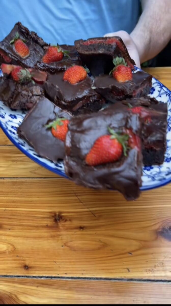 A person holding a plate of chocolate brownies topped with whole strawberries, displayed on a wooden table. Some brownies have bites taken out of them.