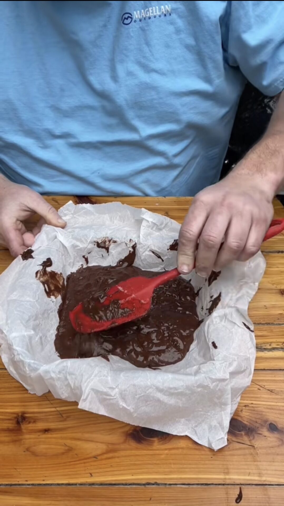 A person in a light blue shirt spreads chocolate batter in a parchment-lined pan with a red spatula on a wooden table.