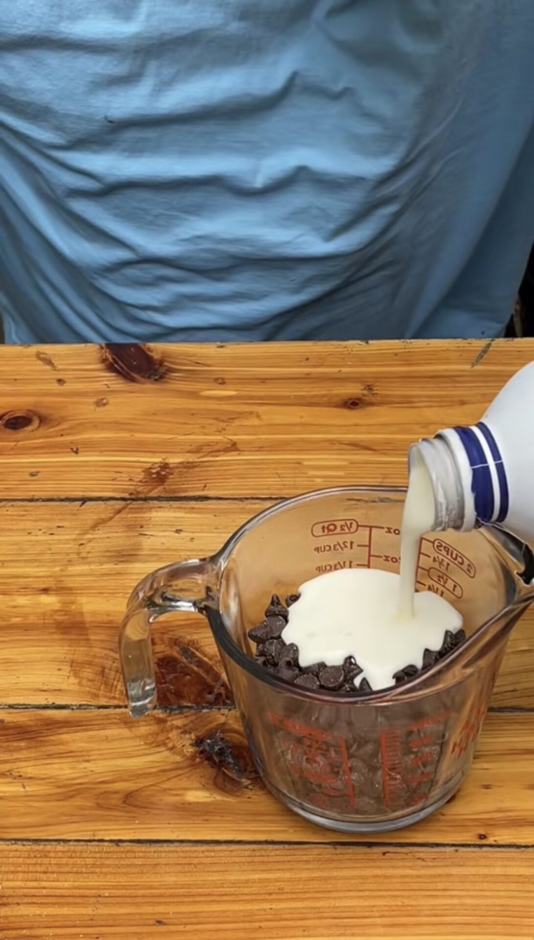 A glass measuring cup filled with chocolate chips is on a wooden surface. Milk is being poured from a bottle into the cup. A person in a light blue shirt stands in the background.