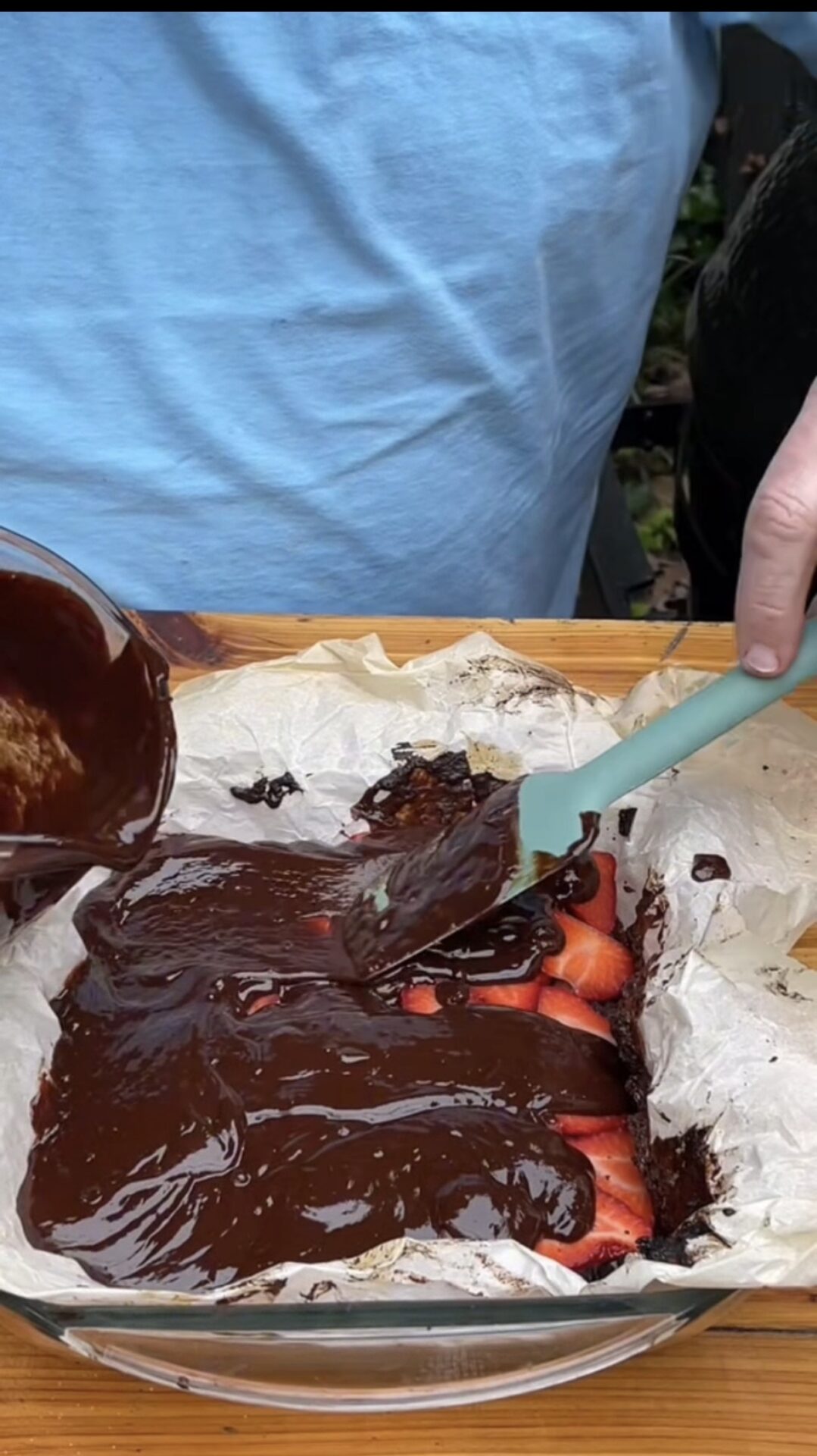 A person pours and spreads melted chocolate over sliced strawberries in a parchment-lined baking dish using a spatula.