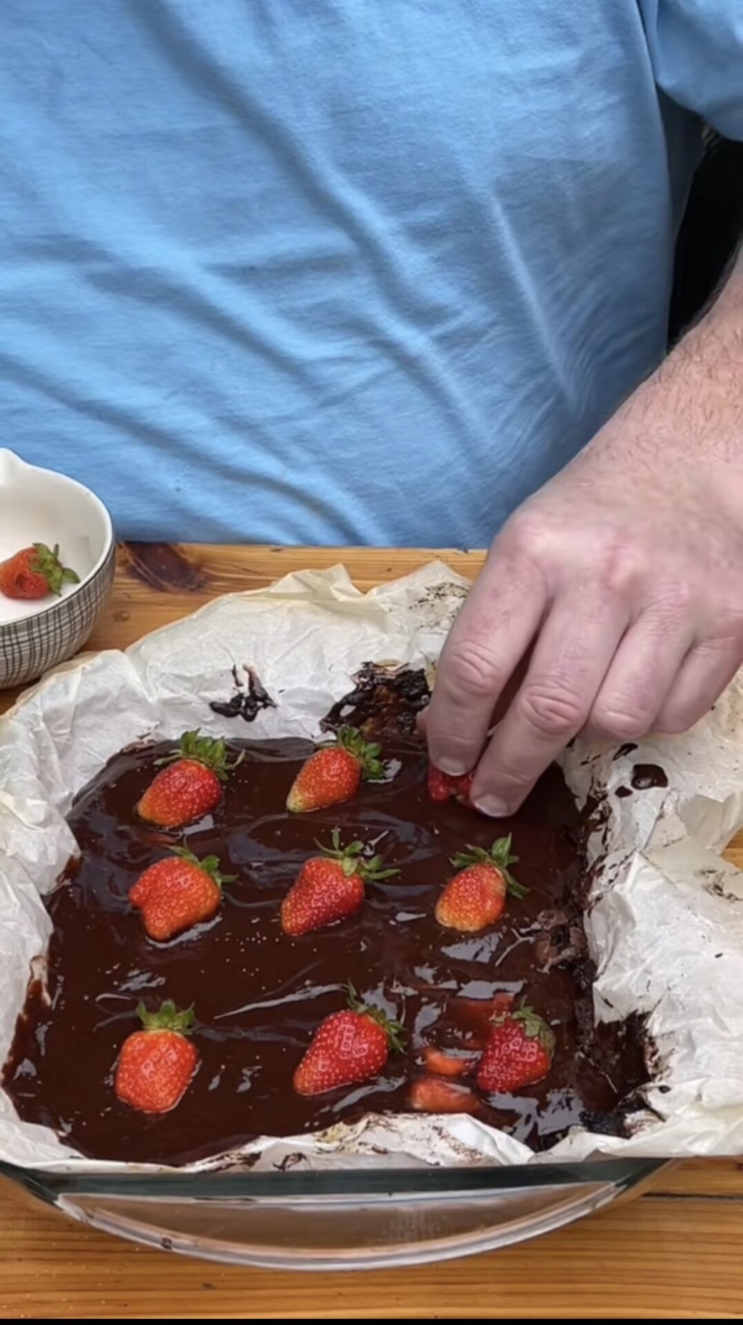 A person wearing a light blue shirt places a strawberry onto a tray of chocolate-covered dessert lined with parchment paper. Several strawberries are already arranged on the glossy chocolate surface.