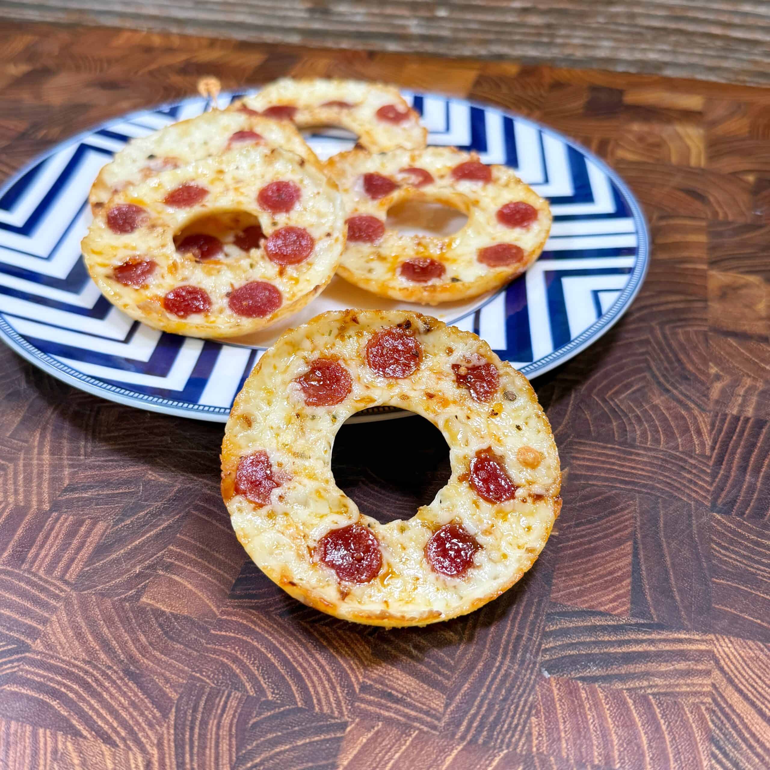 A plate with bagel-shaped mini pizzas topped with small pepperoni slices, placed on a blue and white patterned plate on a wooden surface.