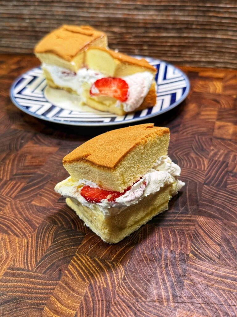 Two slices of sponge cake filled with whipped cream and strawberries, one placed on a wooden surface in the foreground and another on a patterned plate in the background.