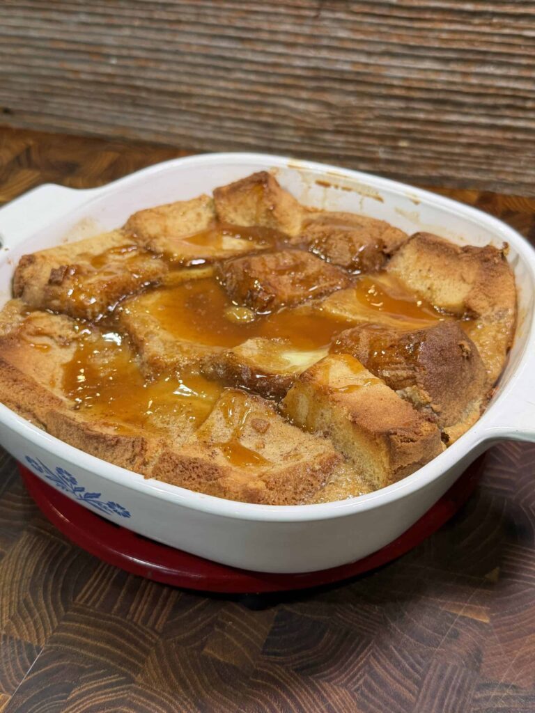 A white ceramic baking dish filled with golden-brown bread pudding, drizzled with caramel sauce, sits on a wooden surface. The dessert appears warm and freshly baked.