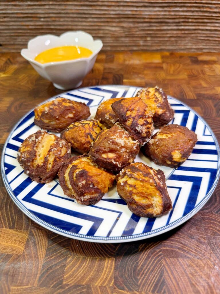 A plate of soft pretzel bites with a golden-brown crust is arranged on a patterned blue and white plate. A small bowl of yellow dipping sauce sits in the background on a wooden surface.