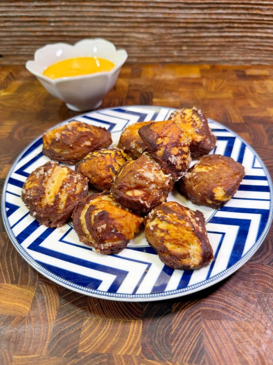 A plate of soft pretzel bites with a golden-brown crust is arranged on a patterned blue and white plate. A small bowl of yellow dipping sauce sits in the background on a wooden surface.