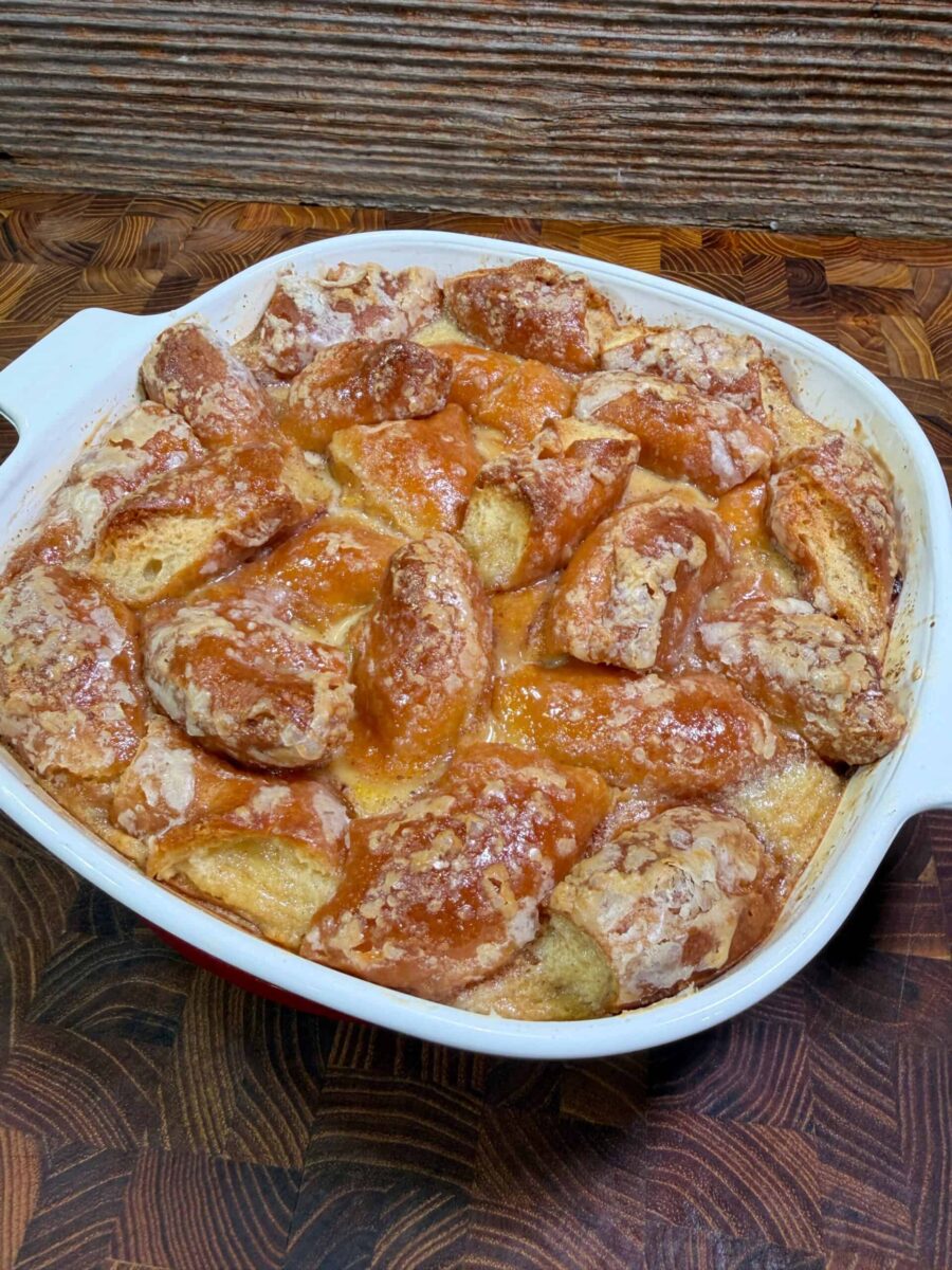 A baked casserole dish, inspired by Krispy Kreme Donut Bread Pudding – Quick Dessert Recipe, features golden-brown, flaky biscuit pieces covered in a shiny glaze, sitting on a wooden surface with a rustic background.