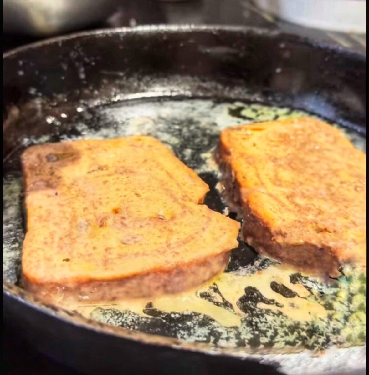 Two slices of bread are being cooked in a black skillet with melted butter, likely to make French toast. The bread is coated in an egg mixture and is browning on the pan.