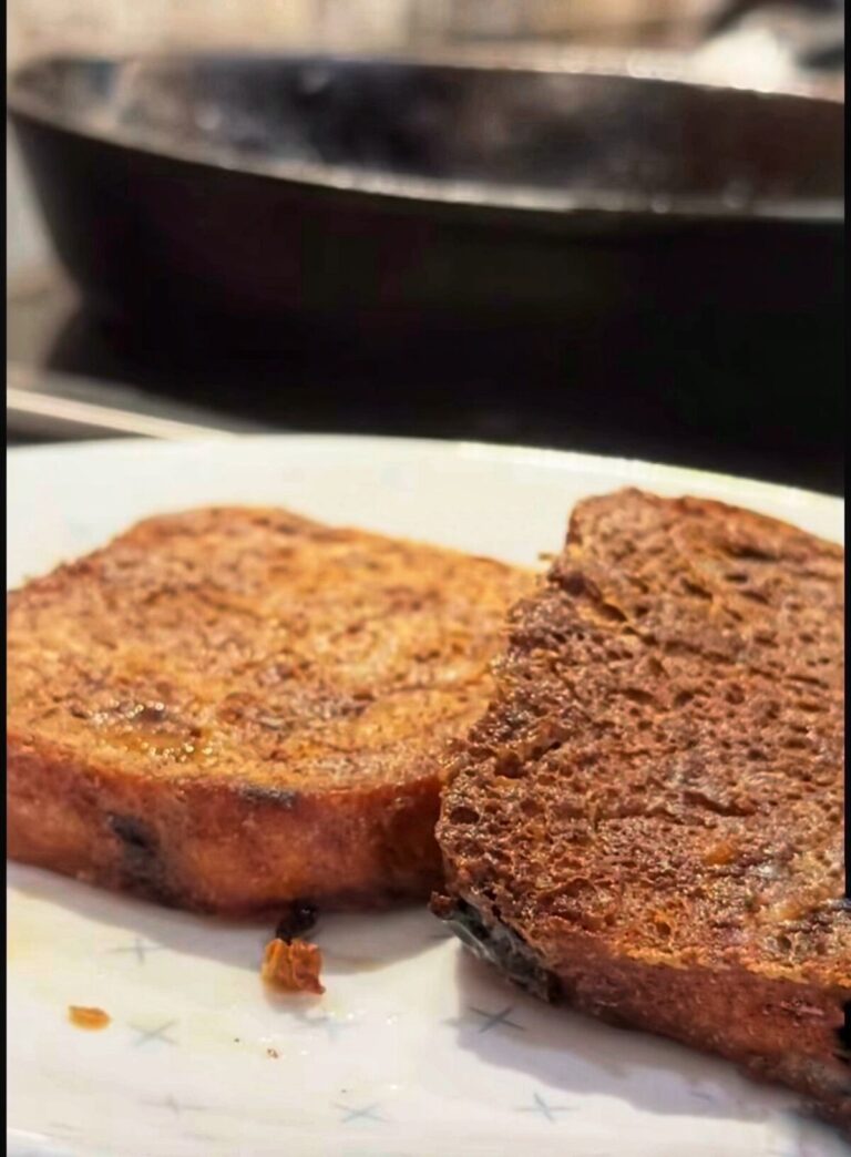 Two slices of cooked French toast rest on a white plate, with a blurred black frying pan visible in the background. The toast looks golden brown and slightly crisp.