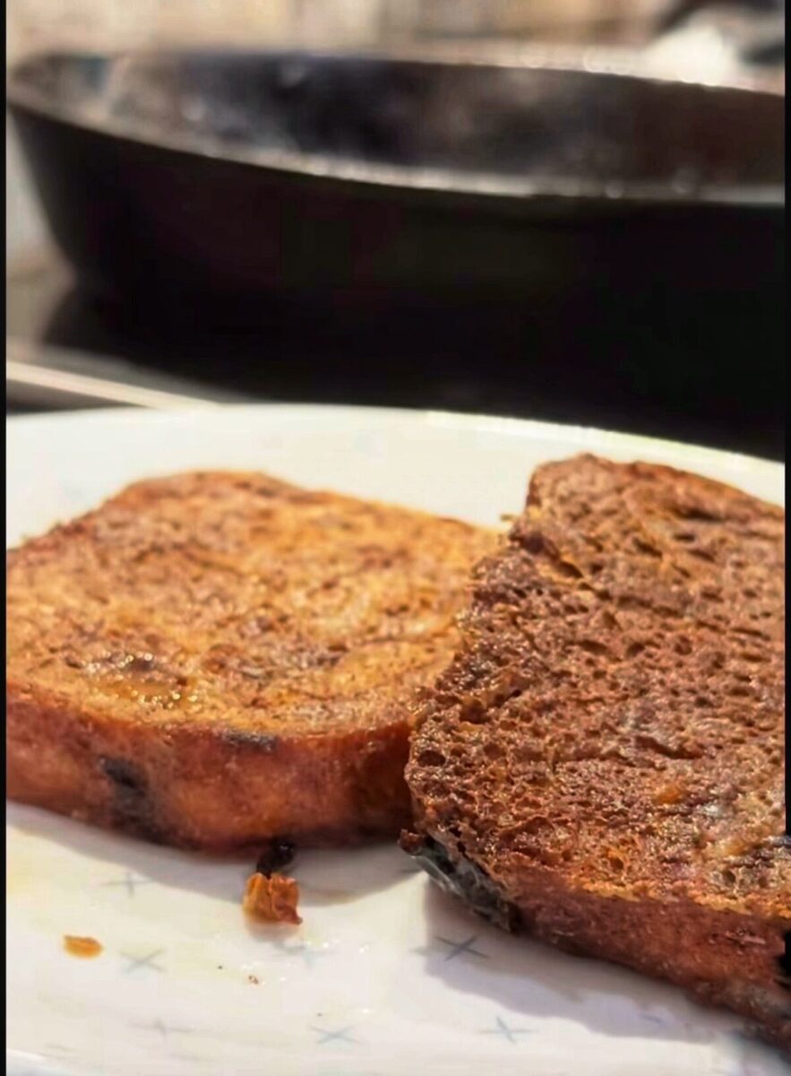 Two slices of cooked French toast rest on a white plate, with a blurred black frying pan visible in the background. The toast looks golden brown and slightly crisp.