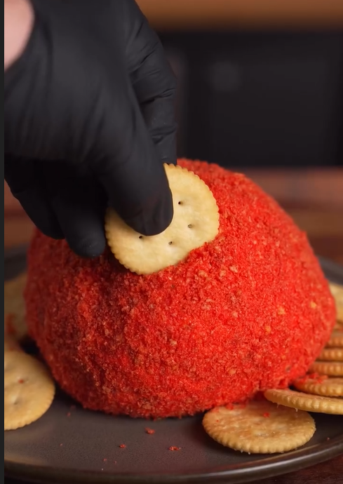 A hand wearing a black glove presses a round cracker into a large, red-coated cheese ball on a plate, surrounded by more crackers.  Recipe for: Spicy Pickleball Cheese Ball