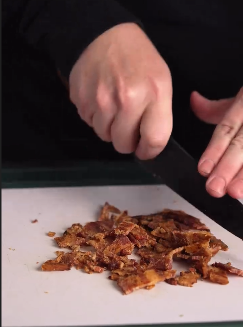 A person chopping cooked bacon into small pieces on a white cutting board with a black knife.