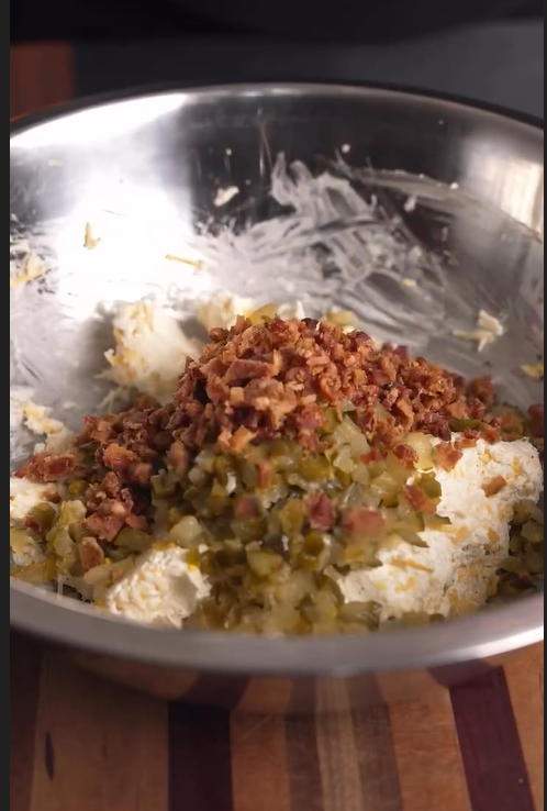 A metal mixing bowl containing cream cheese, chopped pickles, and crumbled bacon sits on a wooden surface, ready to be mixed together.