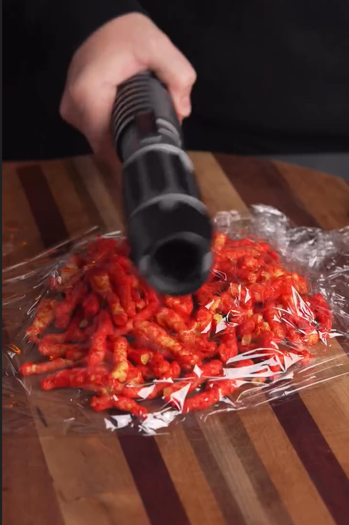 A hand holds a vacuum sealer over a pile of Flamin Hot Cheetos wrapped in plastic wrap on a wooden surface.