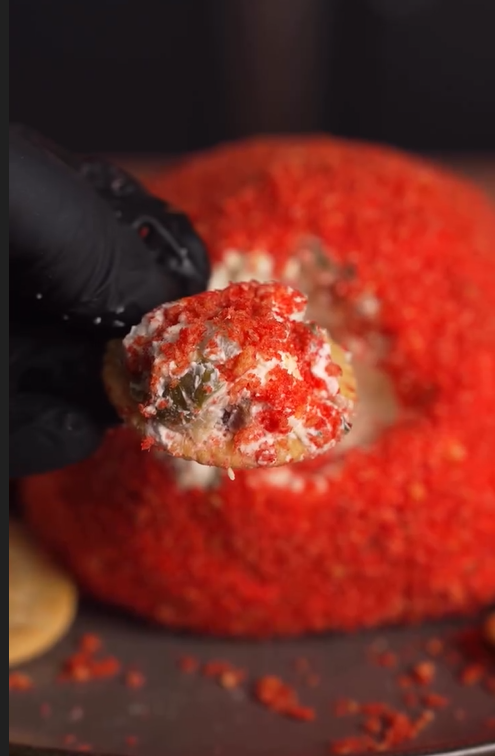 A gloved hand holds a cracker topped with a creamy cheese spread and crushed bright red chips, in front of a large cheese ball coated in the same crushed chips.