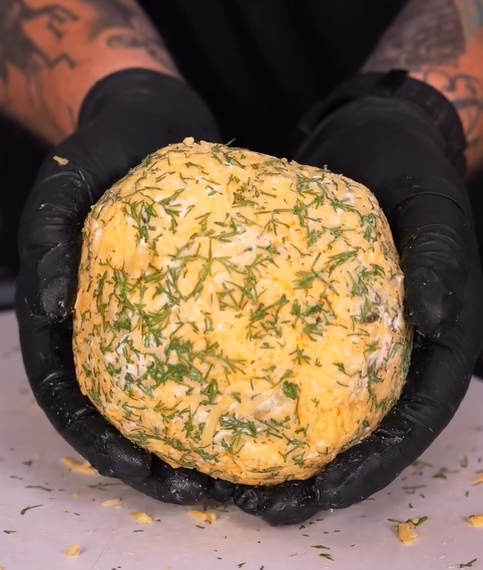 A person wearing black gloves holds a large, round ball of cheese coated with chopped herbs and seasonings. Tattoos are visible on the persons arms in the background.