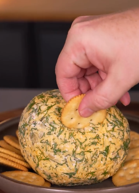 A hand dips a round cracker into a large, herb-covered cheese ball on a plate, with more crackers arranged around the cheese ball.