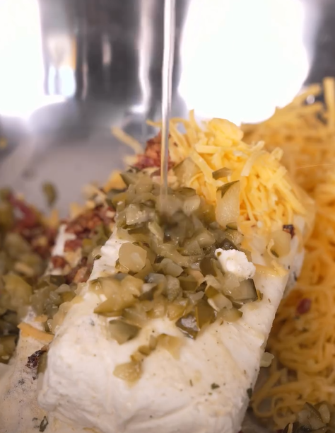 Close-up of cream cheese, shredded cheddar, diced pickles, and seasoning in a metal bowl, with a stream of clear liquid being poured over the ingredients.