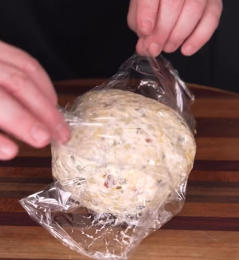 A person’s hands unwrap a ball of cheese mixture with herbs and bits of vegetables from plastic wrap on a wooden cutting board.