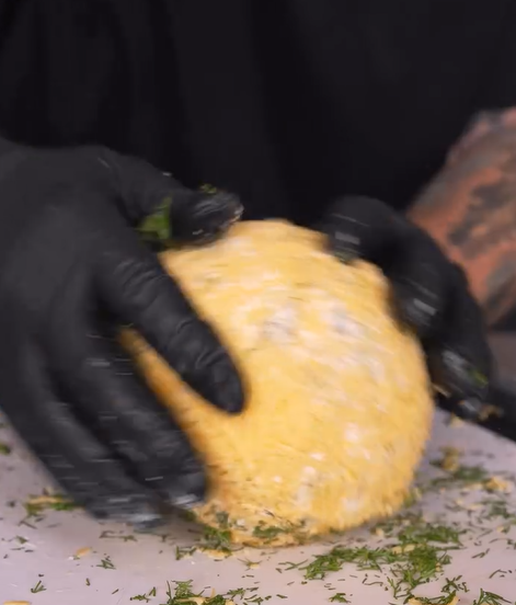 A person wearing black gloves shapes a large, round cheese ball coated with herbs and shredded cheese on a white cutting board.