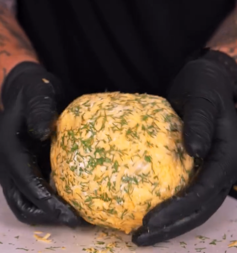 A person wearing black gloves is holding a round ball of cheese coated with herbs and seasonings on a white surface.