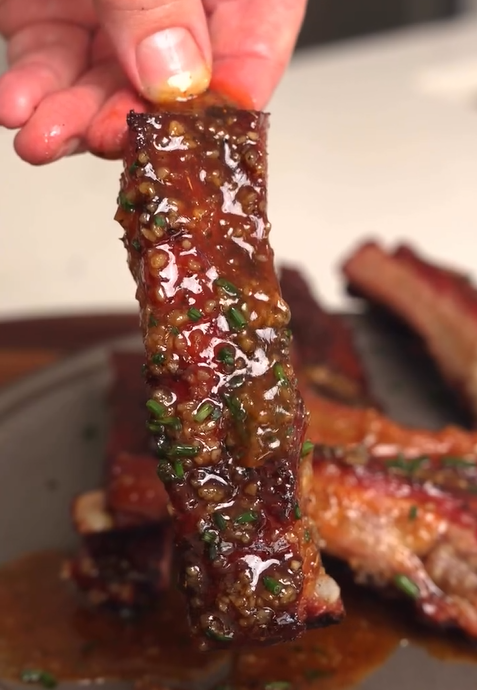 A close-up of a hand holding a glazed, saucy rib topped with chopped herbs over a plate with more ribs in the background. The rib looks tender and caramelized with a glossy sauce.